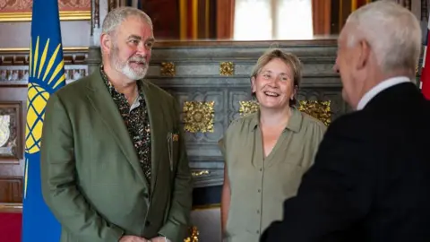 House of Commons Terry Wiggins, wearing a green blazer and patterned shirt, faces the camera, as Lindsey Hoyle (seen from behind) talks to him 