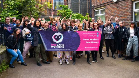 IWGB A group of about 30 casually dressed people standing in the courtyard of an office building. Four people at the front are holding a purple and pink banner with the IWGB union Game Workers' branch logo and the slogan 