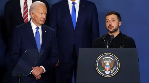 EPA Wearing a blue tie and navy suit, US President Joe Biden stands next to Ukrainian President Volodymyr Zelensky who is speaking at a podium addressing the Nato Summit in Washington in July 2024