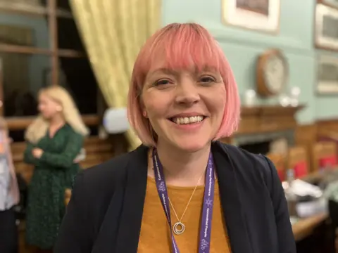 Katie Collier smiling at the camera in the council chamber. She has pink hair, a smart jacket and a purple lanyard.