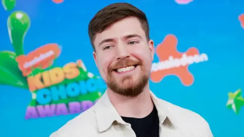 Getty Images Jimmy Donaldson, aka MrBeast, pictured at the Nickelodeon Kids' Choice Awards. MrBeast has short brown hair and a short beard. He wears a white jacket over a black T-shirt and poses in front of a bright blue hoarding with Nickelodeon's orange branding. 