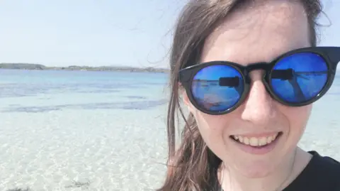 Rebecca Crowe A selfie taken by Rebecca Crowe. She is wearing sunglasses and behind her is some beautiful clear water.