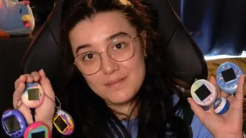 EmmalutionYT A young woman with dark brown hair and glasses holds multiple Tamagotchis in each hand