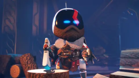 Sony A screenshot of Astro Bot - a white, cute robot with light blue eyes - dressed to resemble Kratos, the protagonist from God of War. Astro has a beard, and wears a leather outfit with lots of buckles and a fur collar. Astro stands next to a chopping block, holding a large axe and is surrounded by logs.