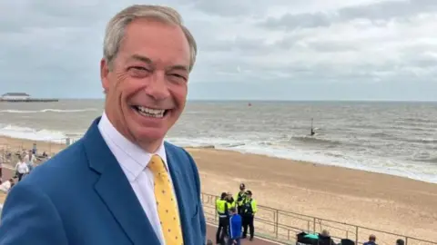 Matt Knight/BBC A smiling Nigel Farage wearing a light blue suit, with white shirt and yellow tie. He is stood above a sandy beach 