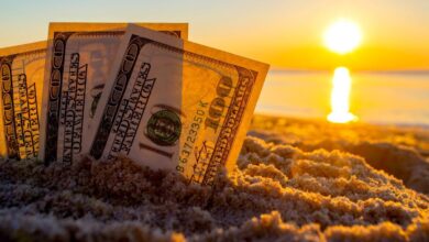 Three one hundred dollar bills stood up and partially buried in the sand, with the sun rising on the horizon.