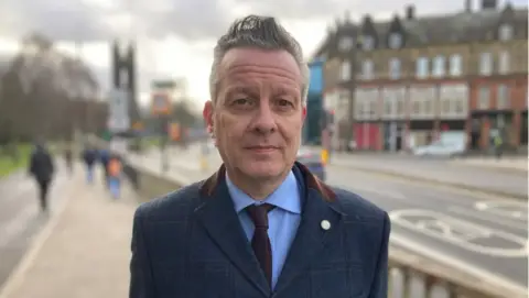 BBC A man with short grey hair is standing on a high street. he is wearing a blue suit and is looking at the camera, expressionless.