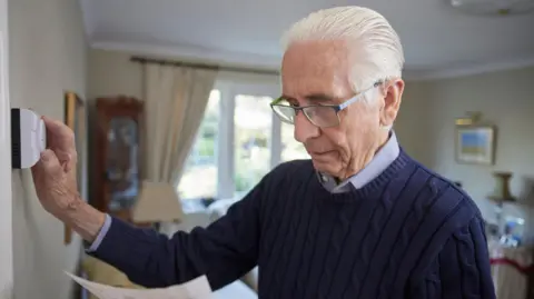 Getty Images Older man at home with a bill in one hand and the other on the thermostat