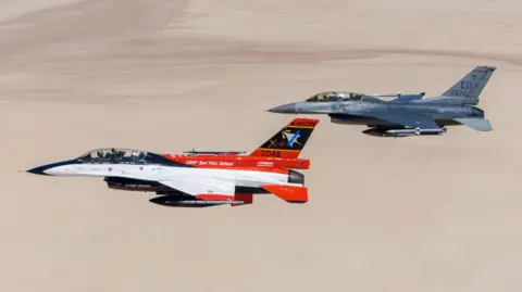 USAF Two US Air Force F-16 combat jets fly side by side over the desert, one white and red, the other combat grey