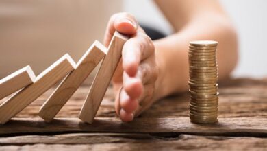 A hand stopping falling dominos from overturning a stock of coins.