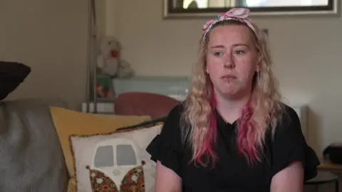 Tamara Beattie wearing a black top and pink bow in her hair, sitting in her home with cushions and a soft duck toy seen behind her