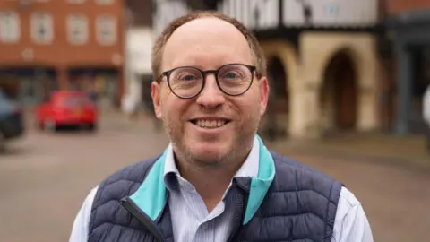 BBC/Simon Dedman Conservative association chairman George Smith smiling at the camera in the Saffron Walden. He has receding brown hair and a beard. He is wearing a blue gilet and a blue-and-white striped shirt.  