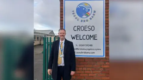 Peter Evans Peter Evans, with short hair and a grey short beard, standing in front of a sign welcoming visitors to Ysgol Bro Banw school, wearing a long black coat, yellow tie and lanyard