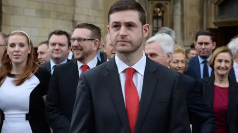 PA Jim McMahon, who has short dark hair and dark stubble, wears a suit and red tie. He is standing in the centre of the frame with a group of politicians and Labour supporters, including Angela Rayner, pictured behind him.