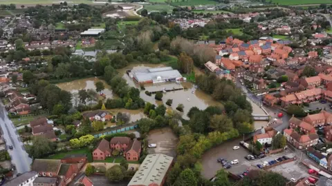 TheDroneMan.net A drone view of a flooded neighbourhood in Horncastle. Brown water covers fields and surrounds buildings, with trees rising above the water.