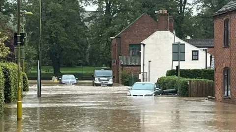 Brown floodwater covers a street in Horncastle, Lincolnshire. Two cars and a taxi and partially submerged. To the right, the water laps against red-brick and white houses to the right. To the left, hedges rise above the water.