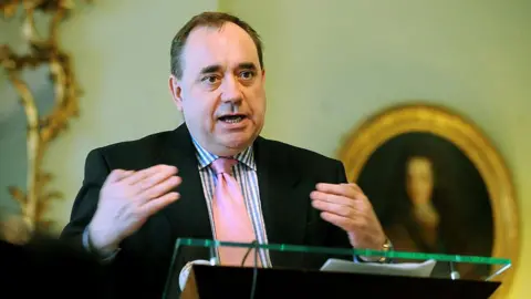 Getty Images Alex Salmond speaking in Bute House in 2008. He is standing at a glass podium, wearing a black suit, checked shirt and pink tie. A portait in an oval gold frame hangs on the wall behind him.