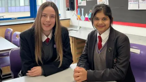 BBC/BRANWEN JEFFREYS Emilia and Dewmi are sitting on purple seats at a white table in a classroom. Emilia is smiling with long brown hair and is wearing her school uniform with a red tie, a grey jumper and black blazer. Demwi is also smiling and her black hair is tied back in a bun. 