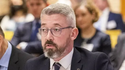 PA Media Niall Ó Donnghaile has grey hair and grey glasses.
He also has a beard that is predominantly grey. 
He is wearing a navy jacket, white shirt and navy tie.