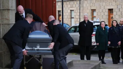 PA Media Four men carry a grey coffin up a few steps through a doorway on the left of the photo. On the right klenny Mackaskill and two women are standing. All are dressed formally in black.