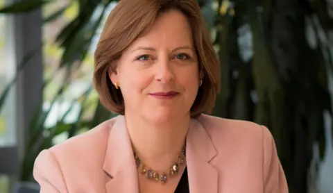 Ofcom Chief Executive of Ofcom Dame Melanie Dawes smiles while looking at the camera and wearing a pink suit with a plant and a window out of focus in the background