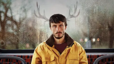 Netflix Promotional image for Netflix show Baby Reindeer, featuring actor and comedian Richard Gadd sitting on the back seat of a bus with antlers drawn in condensation on the window behind him.