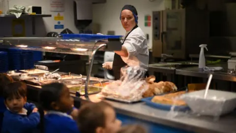 Getty Images A canteen worker spoons food onto a blue tray. She wears gloves, a white top and dark apron and hat. The food is in trays under a hot lamp.