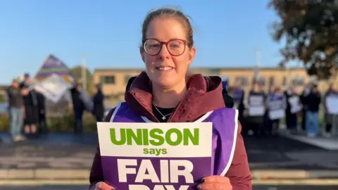 Striking worker Catherine Maciver, wearing large-rimmed glasses with her dark hair tied back, is pictured on a picket line holding a poster which says Unison says fair pay now