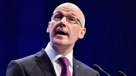 Getty Images John Swinney, in a black suit and purple tie wearing glasses, is mid-speech. He stands in front of a microphone with a dark background.