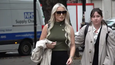 PA Media Victoria Thomas Bowen arriving at Westminster Magistrates' Court in London. She has long blonde hair, and is large black wearing sunglasses, a green sleeveless polo neck top and black skirt. She is carrying a light coloured coat. 