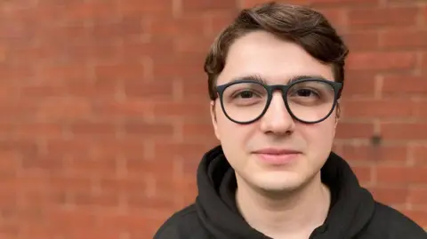 Nathan Standley / BBC Mirzan looks directly into the camera in a close-cropped shot of his head and shoulders. He is wearing glasses and has medium-length wavy brown hair. He is wearing a dark coloured hoodie and stands in front of a blurred brick wall.