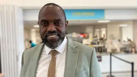 Nathan Standley / BBC Marvin Charles looks into the camera. He is stood in the canteen at his school, in front of the large serving hatch where his pupils have just been in for lunch. He is smiling and wearing a light green suit over a white shirt, with a yellow and brown tie.