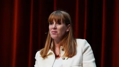 Getty Images Angela Rayner wearing a white jacket sitting down against a red curtain backdrop