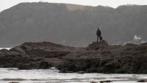 Man stands alone on a rocky outcrop above Cornish shoreline