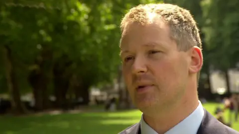 Neil O'Brien head shot, with a leafy background of trees and grass