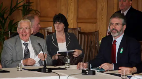 PA Media Ian Paisley and Gerry Adams sitting on two sides at the corner of a table with a white tablecloth and microphones on it.