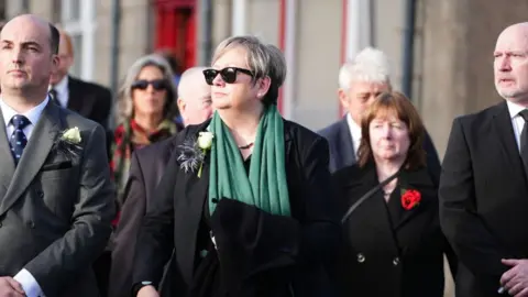 PA Media Joanna Cherry, wearing a dark suit, green scarf and dark sunglasses stands among mourners outside a church 