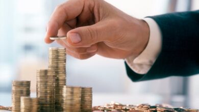 The arm and hand are visible of person wearing professional attire placing a coin onto a stack of other coins.