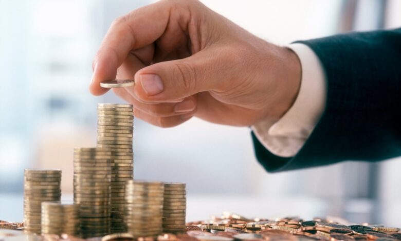 The arm and hand are visible of person wearing professional attire placing a coin onto a stack of other coins.