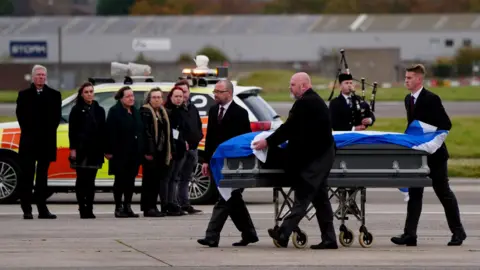 PA Media Alex Salmond's coffin, coverd in a Saltire, is being carried across the tarmac by funeral staff on a trolley. Friends and family are standing in a line in the background