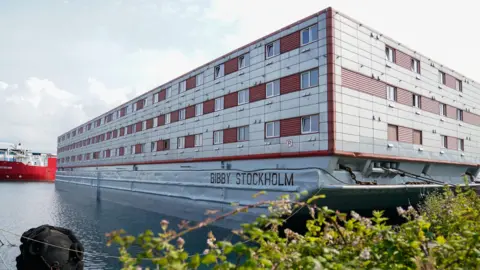 PA Media A view of the Bibby Stockholm, a large silver ship with red windows, being used as accommodation barge at Portland Port in Dorset, which has housed up to 500 asylum seekers at a time. In the foreground is a bush and some water.