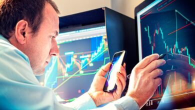 A money manager using a stylus and smartphone to analyze a stock chart displayed on a computer monitor.