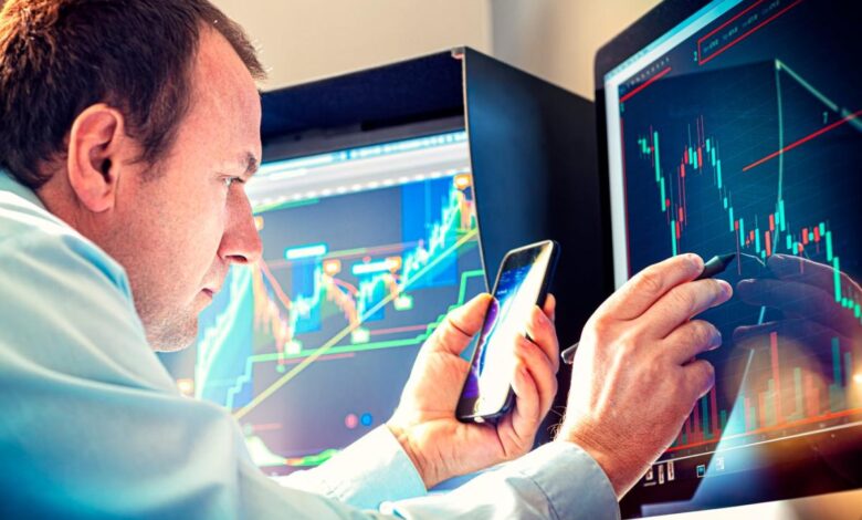 A money manager using a stylus and smartphone to analyze a stock chart displayed on a computer monitor.