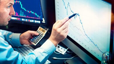 A money manager using a pen and calculator to analyze a stock chart displayed on a computer monitor.