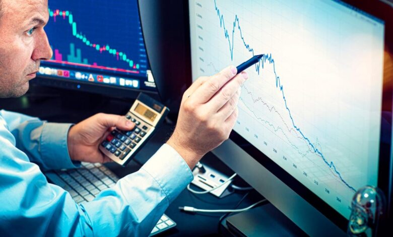 A money manager using a pen and calculator to analyze a stock chart displayed on a computer monitor.