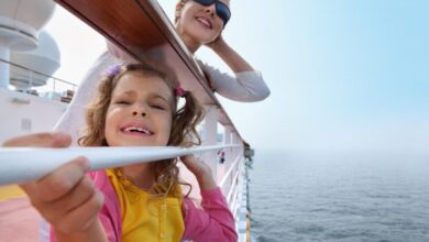 A parent and child smile while holding onto a railing on the deck of a cruise ship.