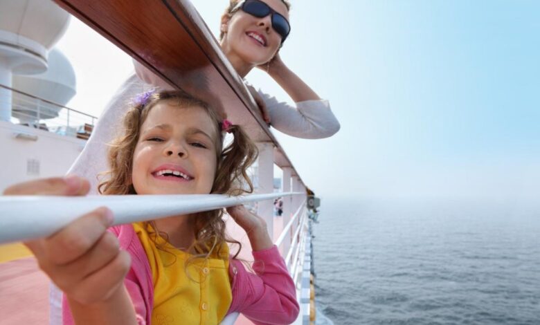 A parent and child smile while holding onto a railing on the deck of a cruise ship.