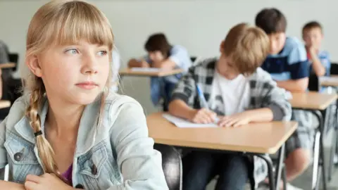 Getty Images A school classroom scene with a blonde girl gazing to the left and three boys sitting and writing at desks in the rows behind her.