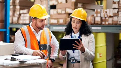 Getty Images Employees wearing hard hats in a warehouse discussing business while looking at an computer tablet