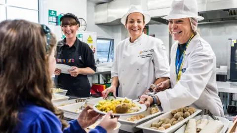 Durham County Council A young school girl is being handed a plate of food from a serving counter by three smiling dinner ladies. She is turned away from the camera but has long brown hair and a blue school jumper. Two of the three dinner ladies wear white overalls and hair nets, while the third has a black apron on.